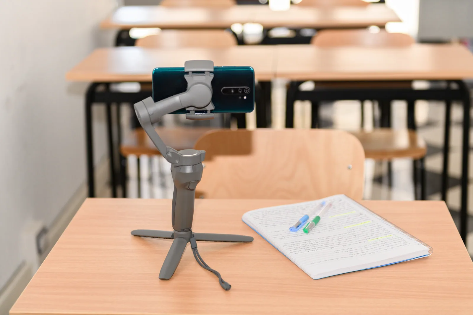 Boş sınıfta tripoda takılı telefon Boş sınıftaki sıranın üzerinde tripod ve sabitleyiciye takılı akıllı telefon ile not kâğıtları; okulda fotoğraf çekimini anlatıyor